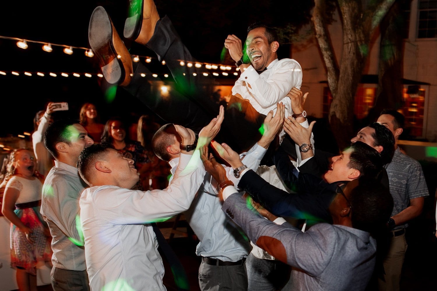 A joyful group of people raises a smiling man in a white shirt into the air during a nighttime celebration under the starlit sky at Leach Botanical Garden.