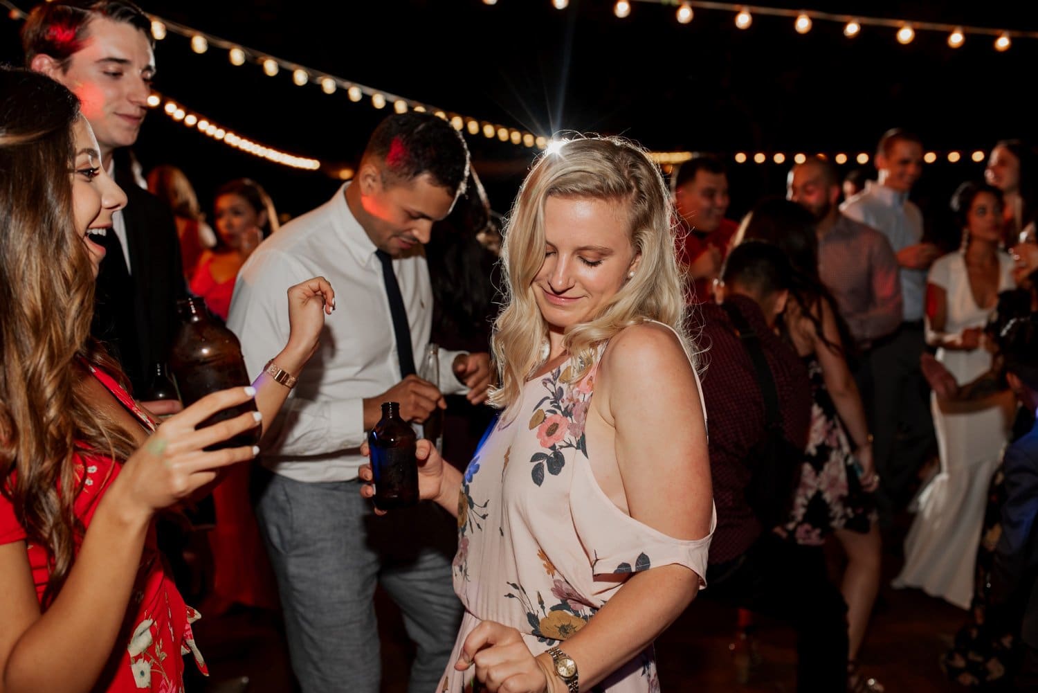 Guests sway and mingle under the stars at a vibrant nighttime celebration, with string lights illuminating the area overhead.
