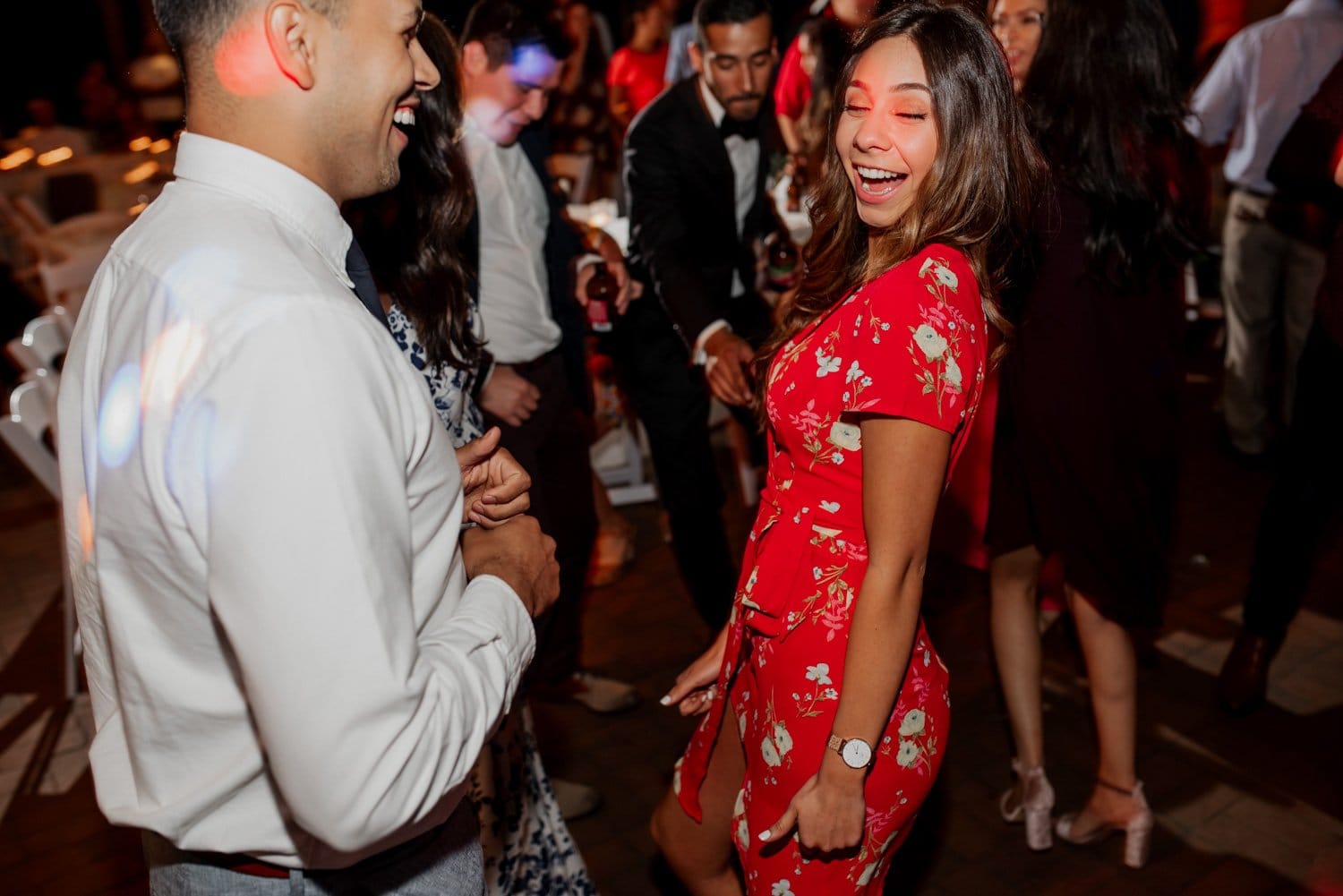 At a lively gathering at Leach Botanical Garden, people are dancing and socializing. A woman in a red floral dress stands out as she smiles brightly.