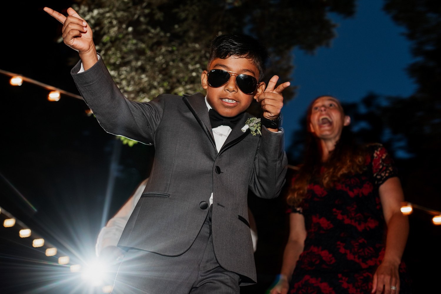 A young boy in a suit and sunglasses dances energetically under the night sky at Leach Botanical Garden, with a woman laughing behind him.