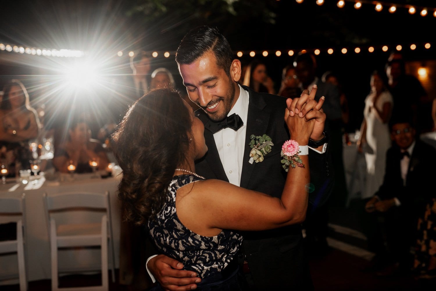 A couple dances closely under twinkling lights at their Leach Botanical Garden wedding, with guests celebrating in the background.