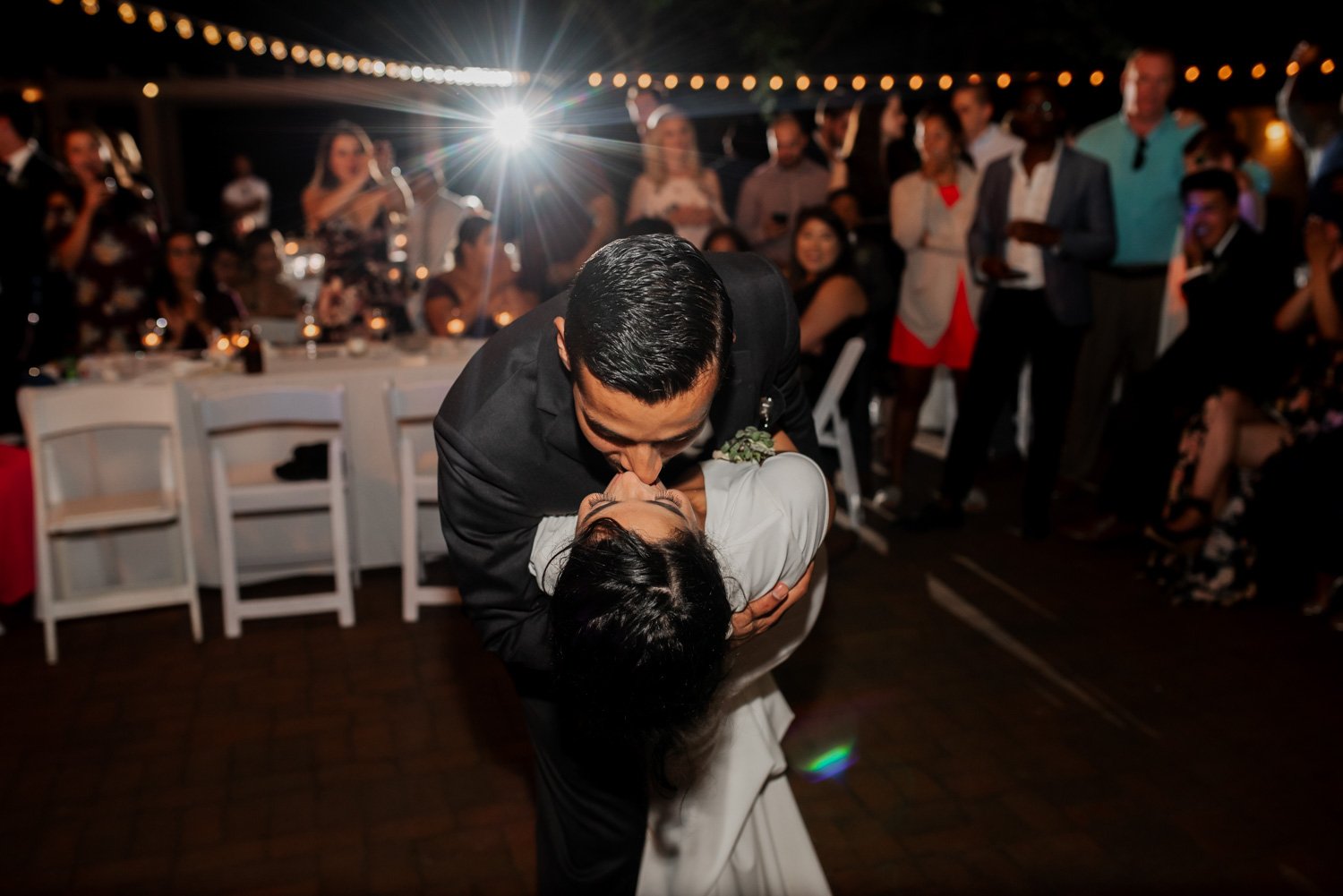 A couple shares a romantic kiss under twinkling string lights at Leach Botanical Garden, with guests smiling around them as a camera flash illuminates the scene.