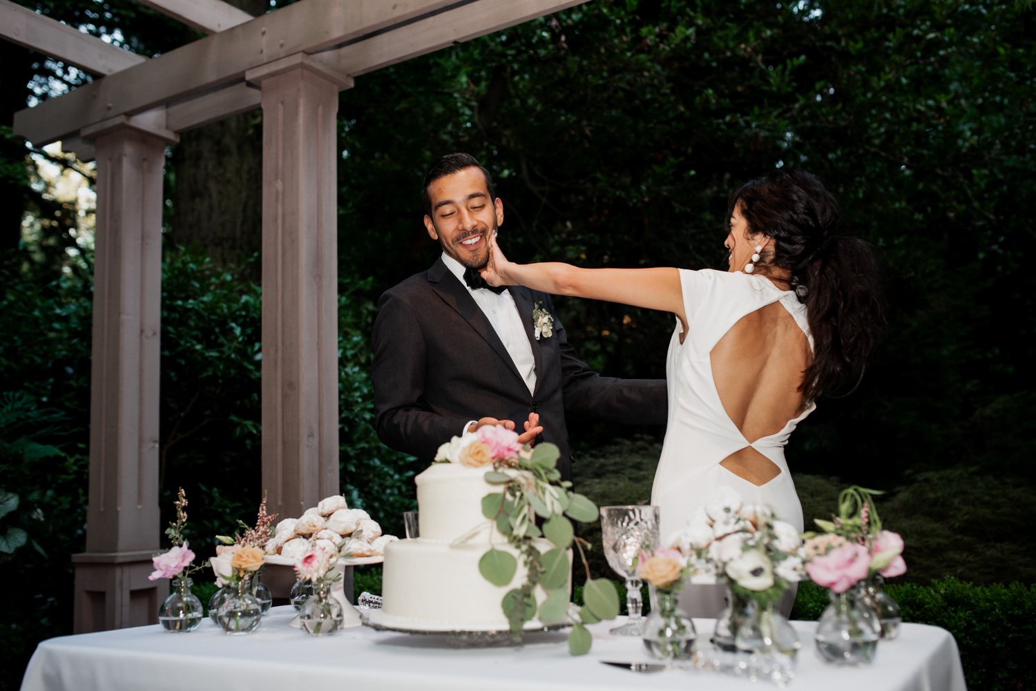 Bride playfully smearing cake on grooms face at outdoor wedding with guests laughing around the decorated cake table.