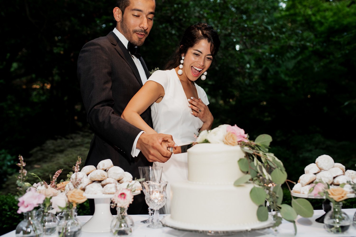 A couple smiling and cutting into a white tiered wedding cake, with elegant flower arrangements and assorted pastries in the background at Leach Botanical Garden.