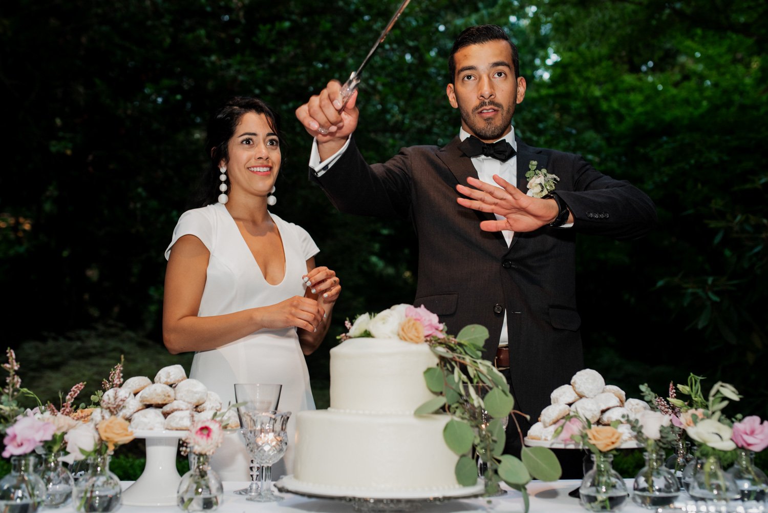 The groom holds a saber high, preparing to cut the wedding cake, while the bride smiles beside him. They are surrounded by flowers and a variety of donuts.