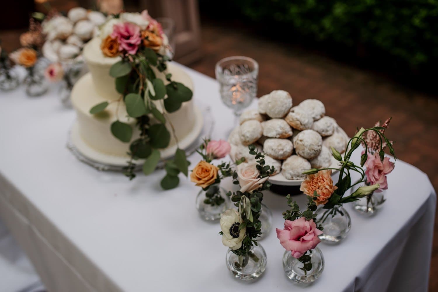 A wedding cake with floral decorations is displayed next to powdered doughnuts. Both are set on a table at Leach Botanical Garden, surrounded by small vases filled with colorful flowers.