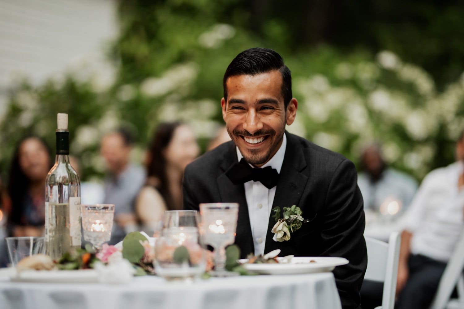 A man in a suit smiles while sitting at a decorated table outdoors, surrounded by bottles and glasses, during a celebration at Leach Botanical Garden.