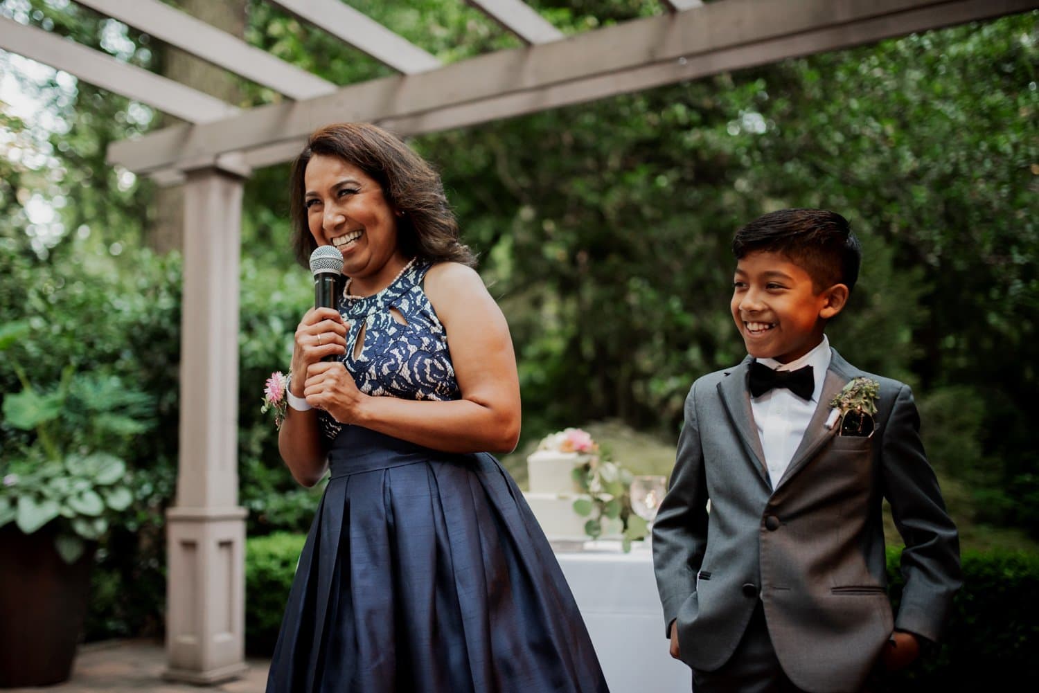 A woman smiling and speaking into a microphone stands next to a boy in a suit, both surrounded by the lush greenery of Leach Botanical Garden, depicting the happiness of a garden wedding.