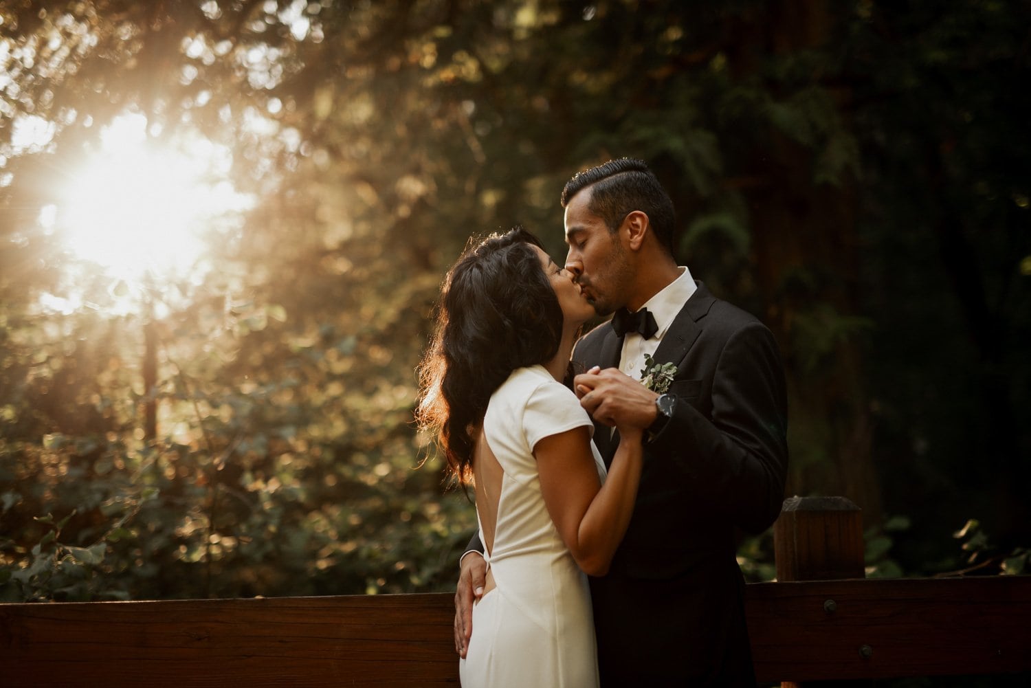 In the Leach Botanical Garden, a couple shares a kiss in a sunlit forest, with the bride in a white dress and the groom in a black suit.