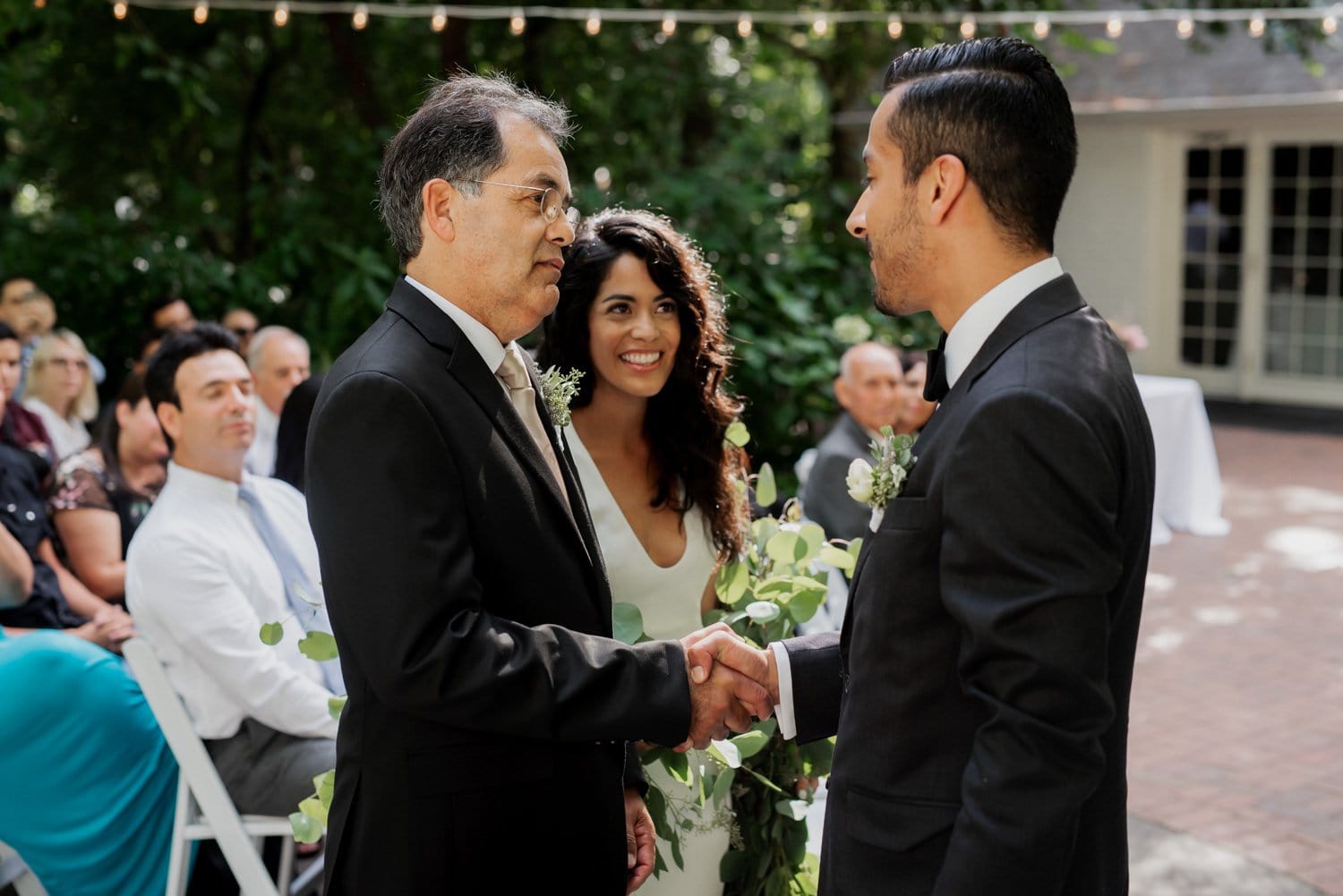 The groom shakes hands with a man while the bride smiles joyfully in an outdoor setting at Leach Botanical Garden.