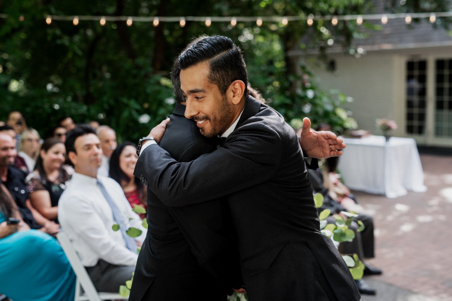 Two men in suits share a warm embrace at a wedding held in Leach Botanical Garden, with seated guests visible in the background and string lights illuminating the scene.