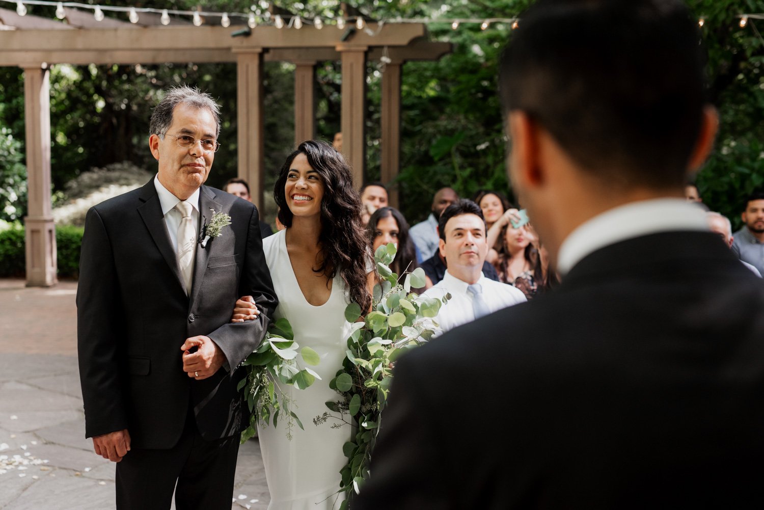 A bride walks down the aisle with an older man at an outdoor wedding ceremony at Leach Botanical Garden, smiling at the groom. The garden is lush and green, providing a beautiful backdrop for the event.