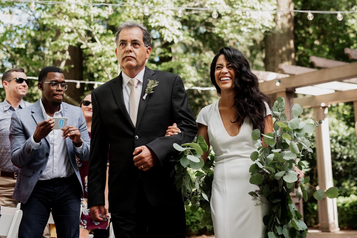 A bride walking down the aisle at Leach Botanical Garden, holding a colorful bouquet and smiling. A man in a suit walks beside her during the outdoor wedding ceremony.