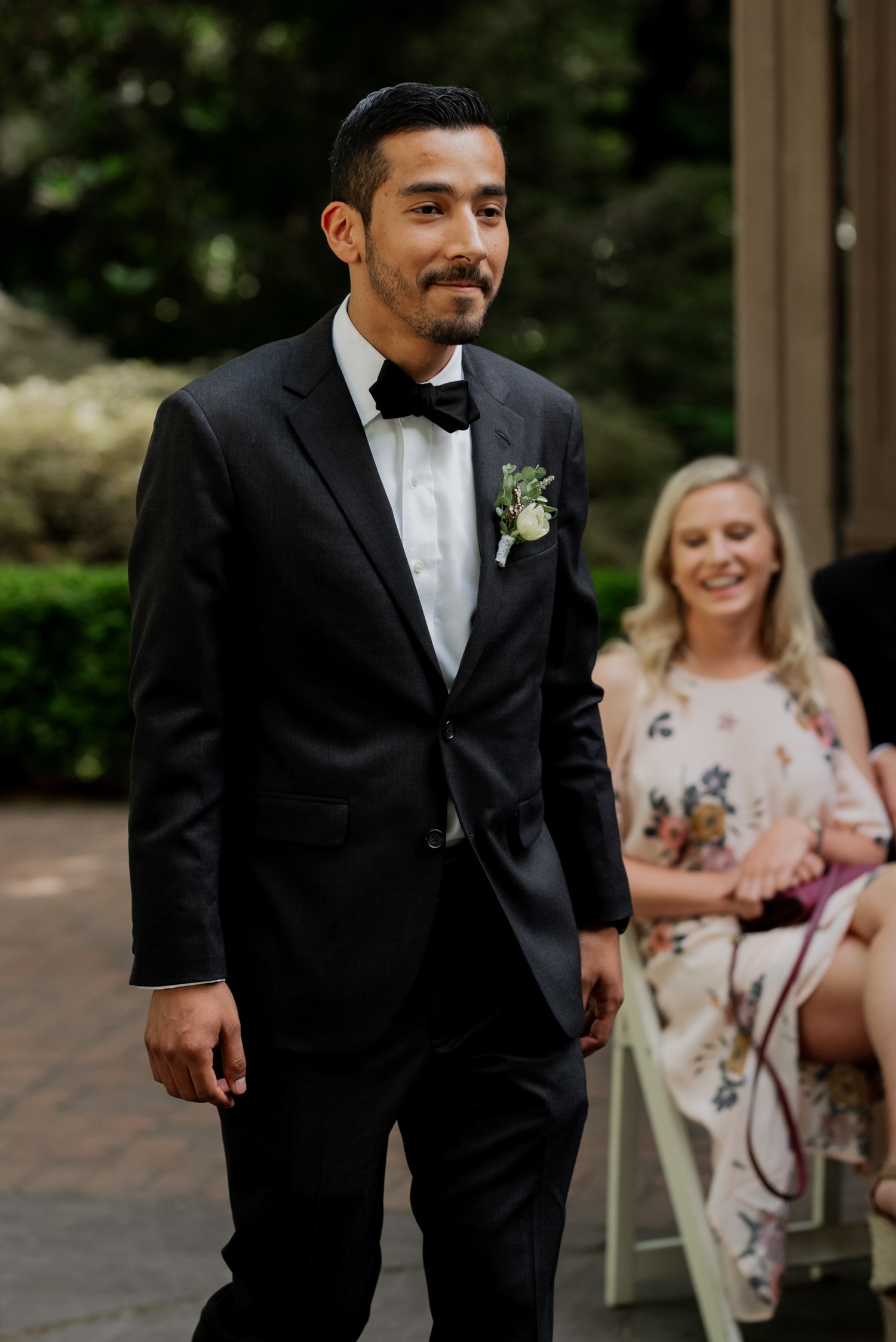 A man in a dark suit with a boutonniere walks past seated guests, surrounded by lush greenery at a Leach Botanical Garden wedding.
