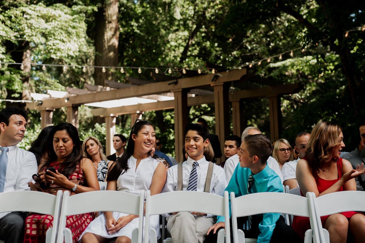 A group of people seated outdoors at a Leach Botanical Garden wedding, engaging in conversation and smiling. The setting is surrounded by trees and illuminated by string lights.