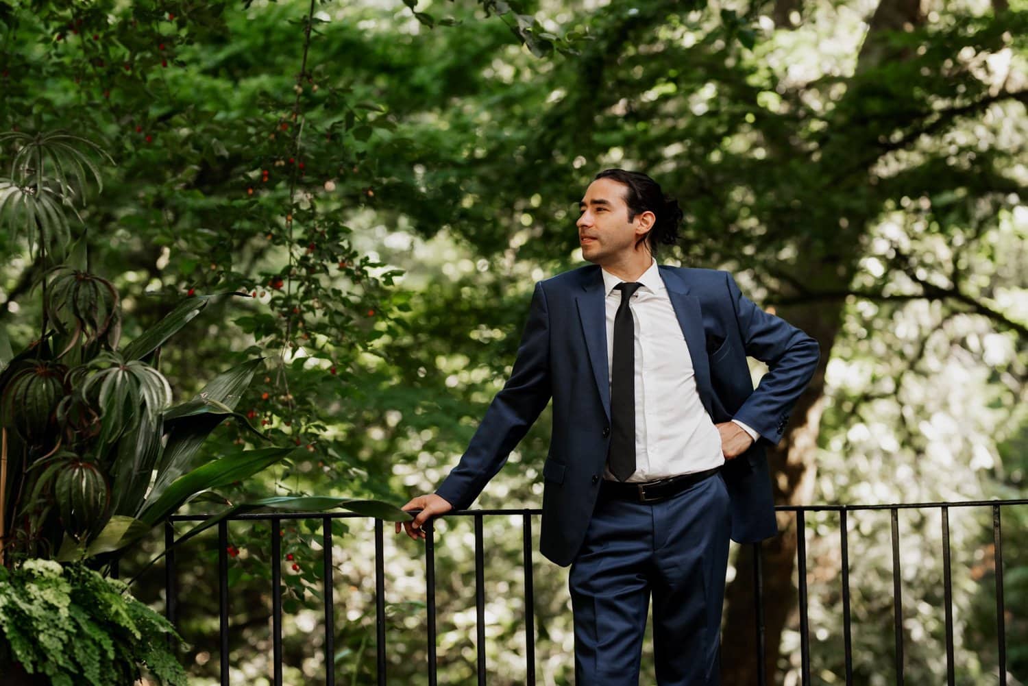 A man in a blue suit stands on a balcony, surrounded by lush green trees.