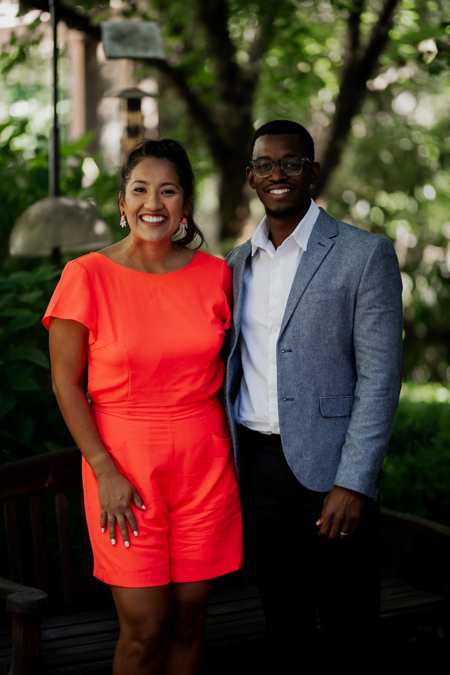 A smiling woman in a bright red outfit stands beside a man in glasses and a blue blazer, surrounded by the greenery of Leach Botanical Garden on their wedding day.