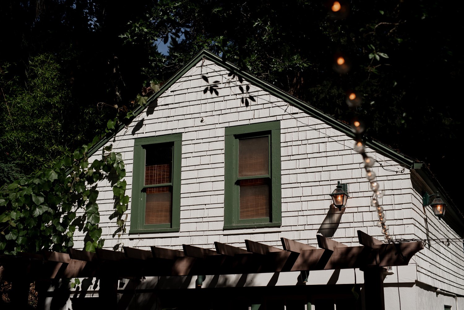 A charming white house with green trim is surrounded by trees. In the foreground, a wooden pergola decorated with string lights is visible.
