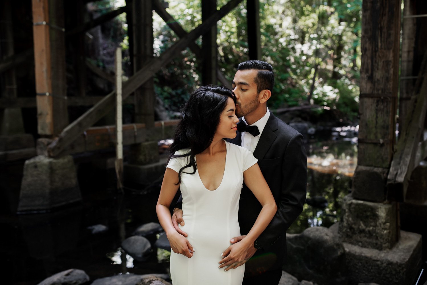 A couple embraces under a wooden bridge next to a stream, surrounded by lush greenery at Leach Botanical Garden during their wedding.