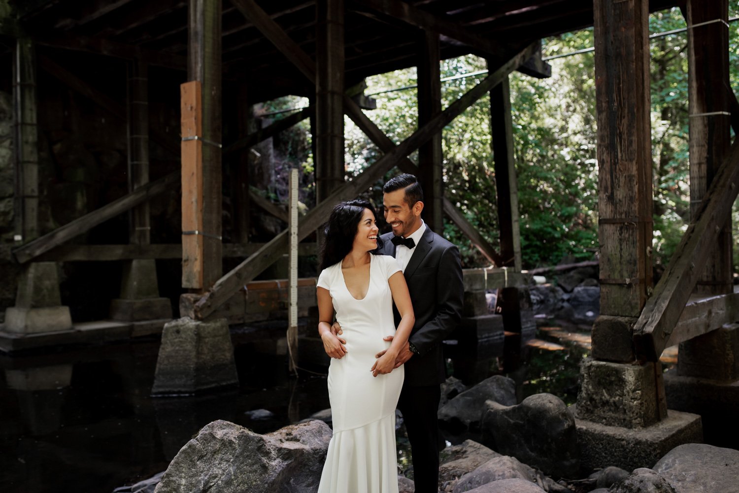 A couple in elegant wedding attire embraces and smiles beneath a wooden bridge near a stream at Leach Botanical Garden.