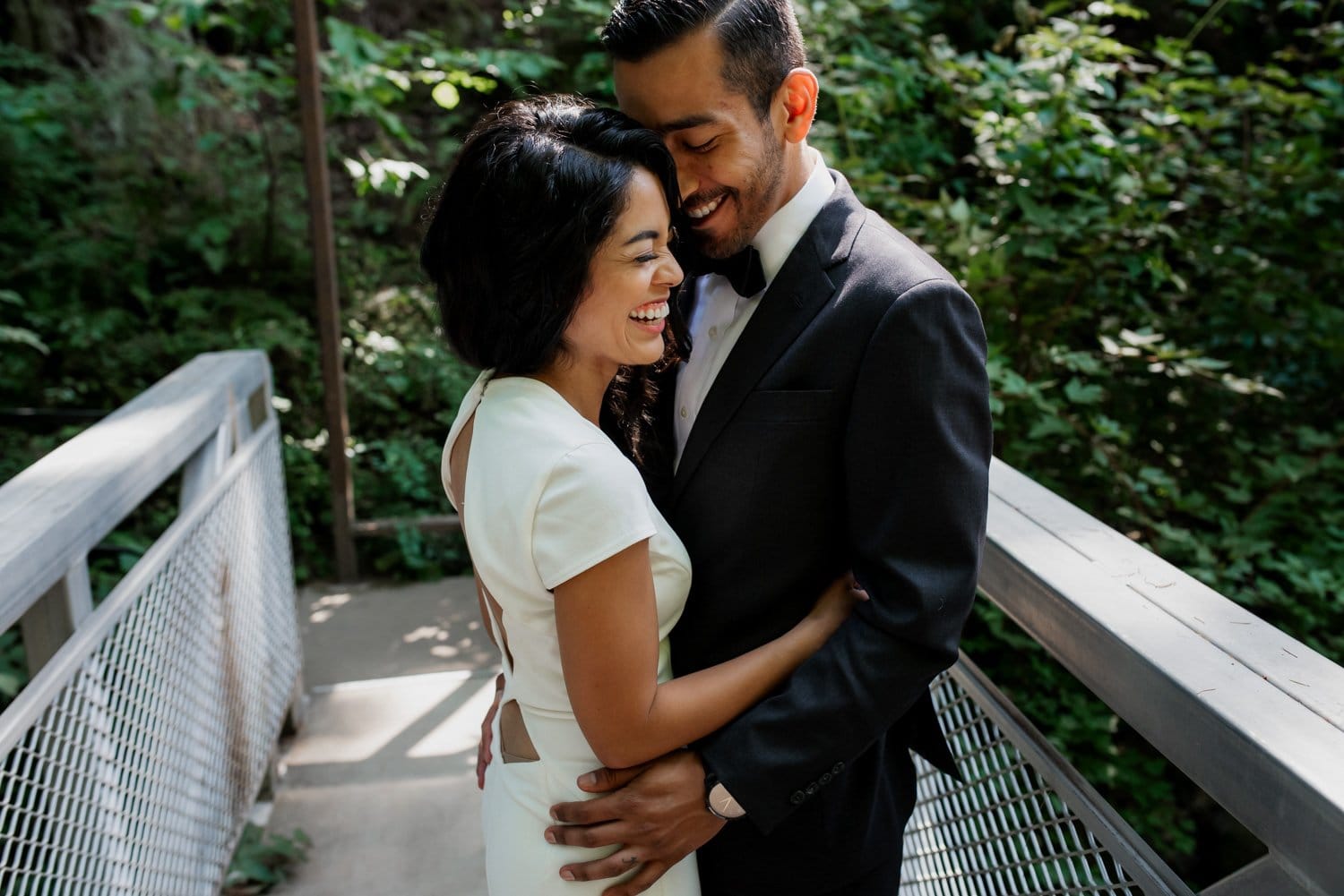 A couple embraces and laughs on a wooden bridge, surrounded by lush green foliage at a botanical garden.