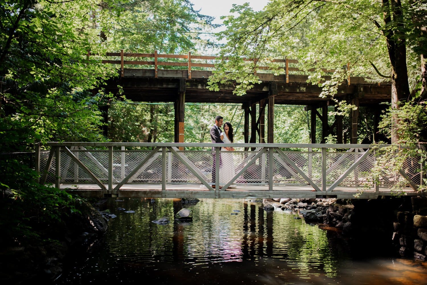 A couple stands on a bridge over a calm stream in a forested area, with sunlight filtering through the trees.