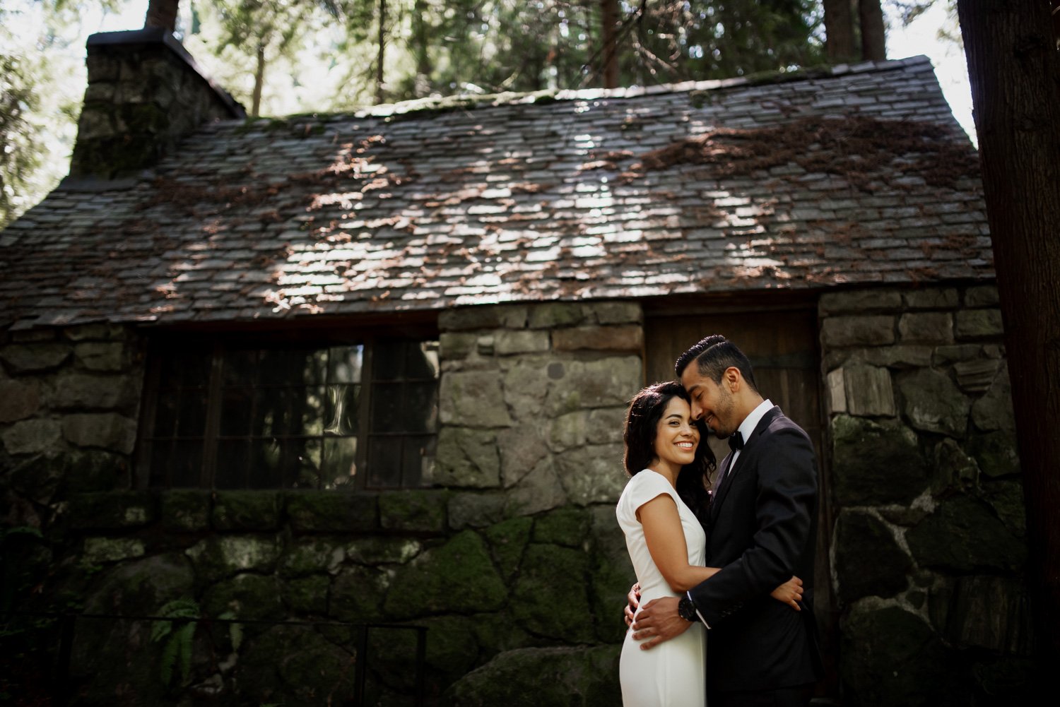 The bride and groom embrace in front of a rustic stone cottage, surrounded by the lush greenery of Leach Botanical Garden.