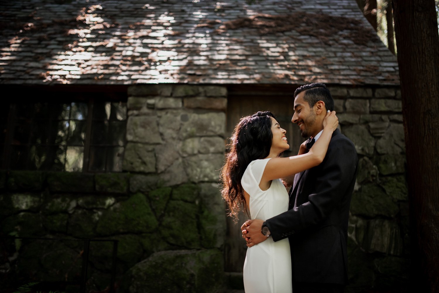 A couple embraces lovingly in front of a rustic stone cabin surrounded by trees.