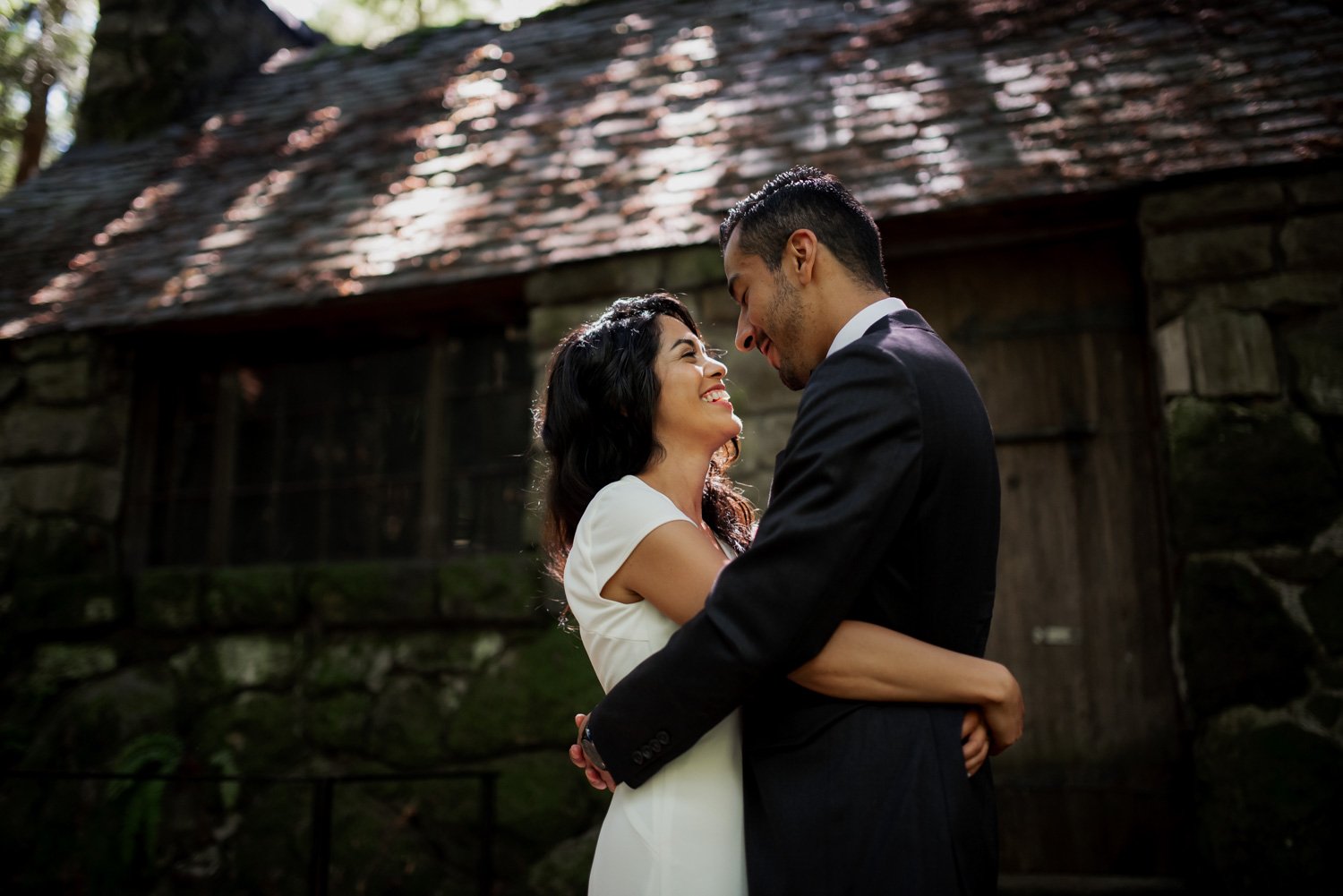 A couple in formal attire embraces and smiles at each other in front of a rustic cabin, celebrating their wedding at Leach Botanical Garden.