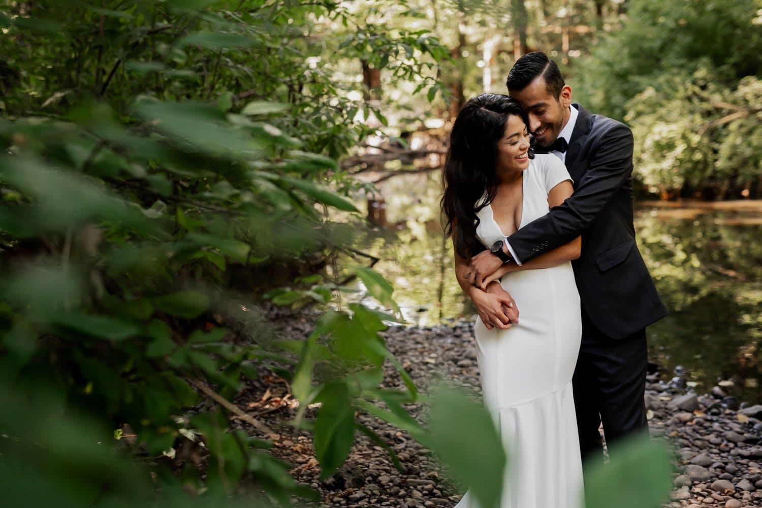 A couple embraces by a forest stream at Leach Botanical Garden, the woman wearing a white dress and the man in a black suit, with lush greenery surrounding them.