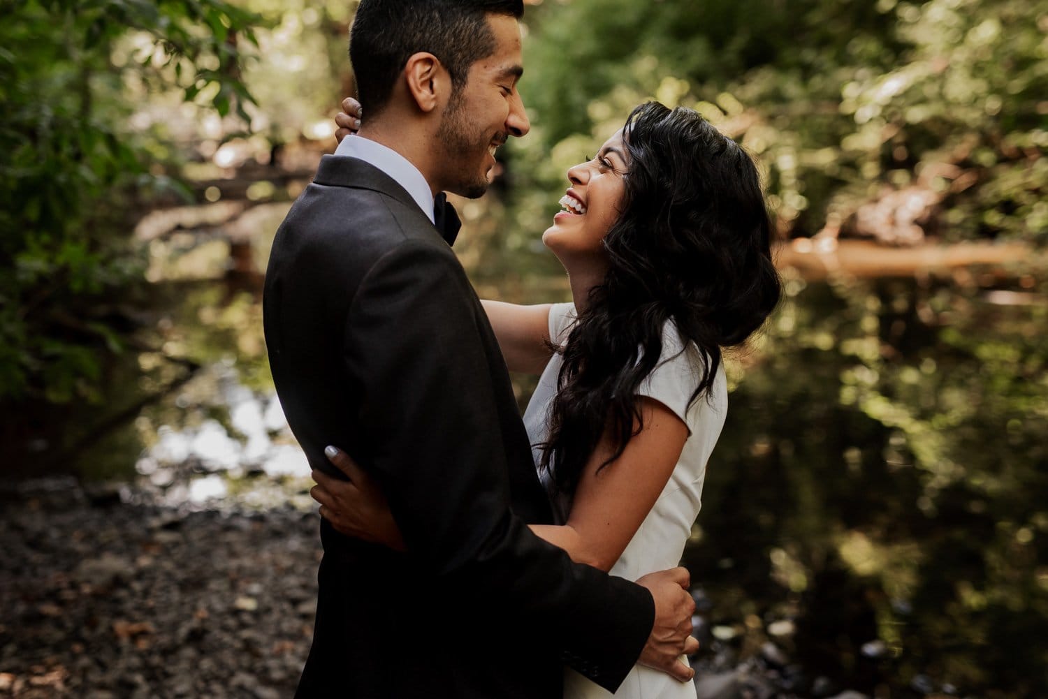 A couple in formal attire embraces and laughs by a forested riverside, with tall trees and calm water in the background.