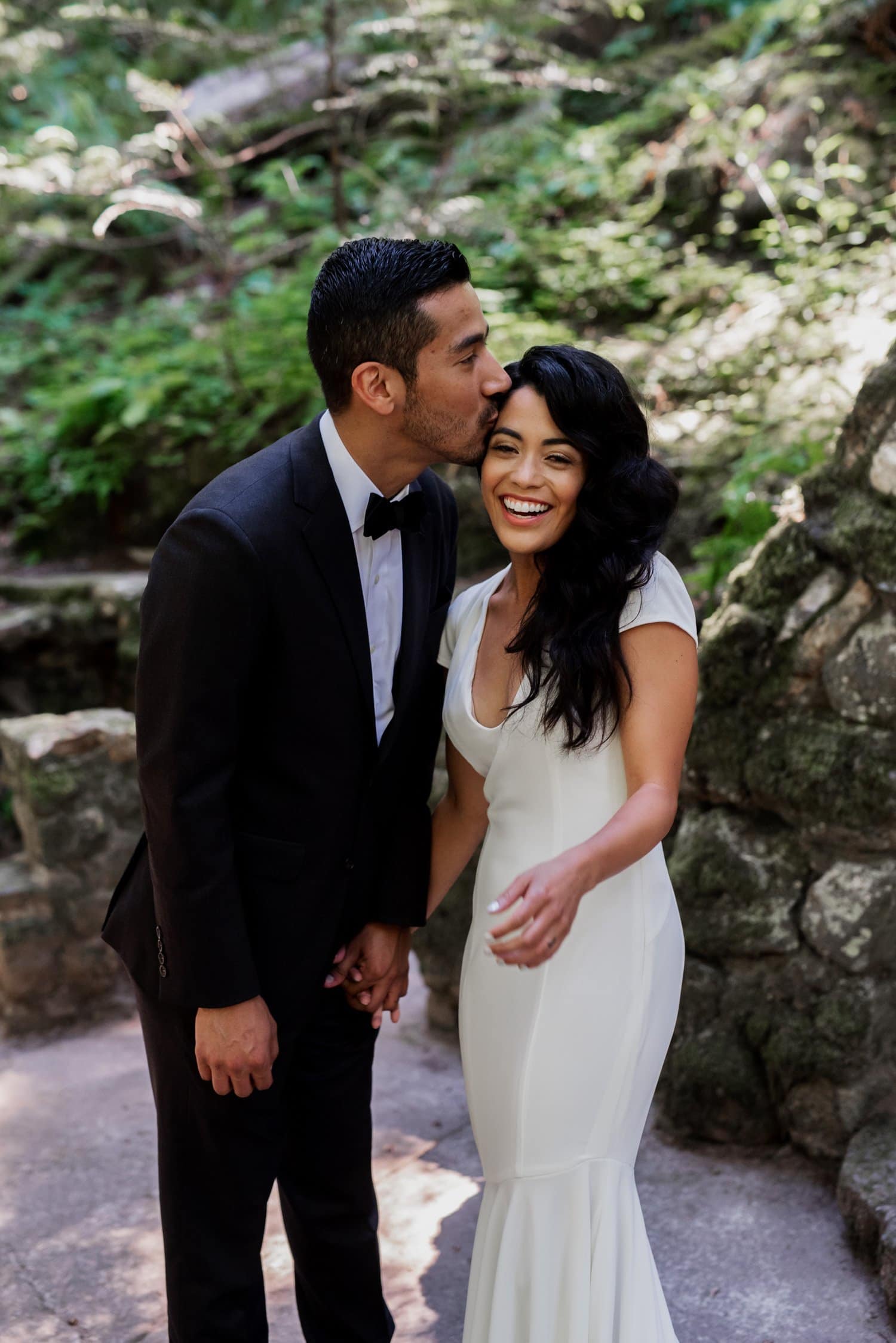 A groom gently kisses his smiling bride on the forehead in a tranquil, wooded setting at Leach Botanical Garden.