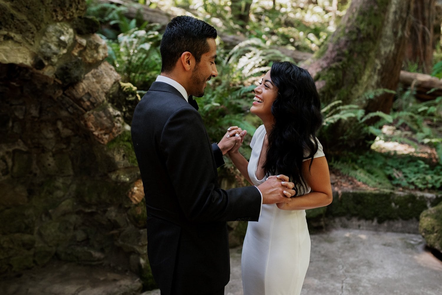 A smiling couple holds hands in a forest setting at Leach Botanical Garden, elegantly dressed in formal attire, on their wedding day.