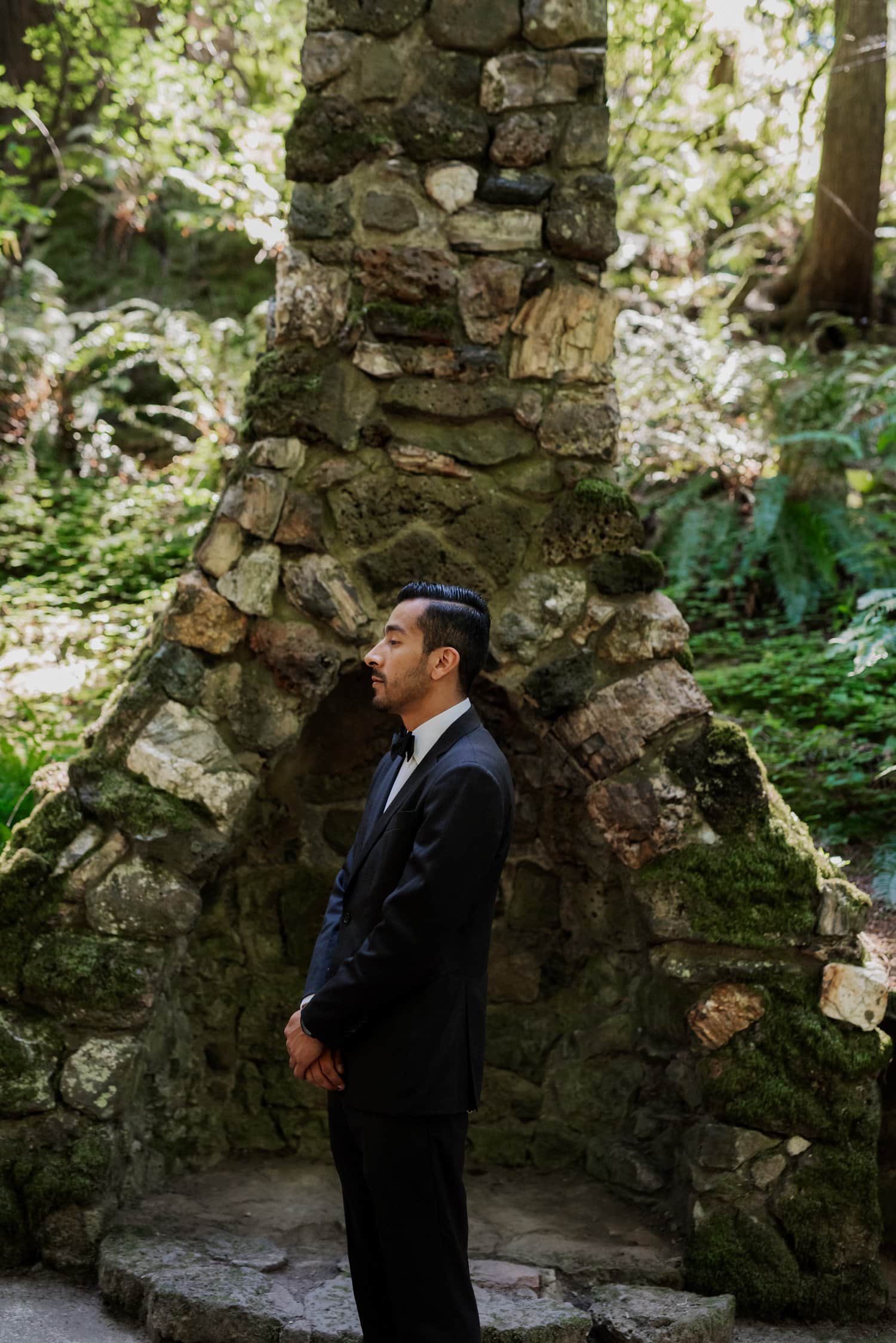 A man in a suit stands by a moss-covered stone structure in a forest setting.