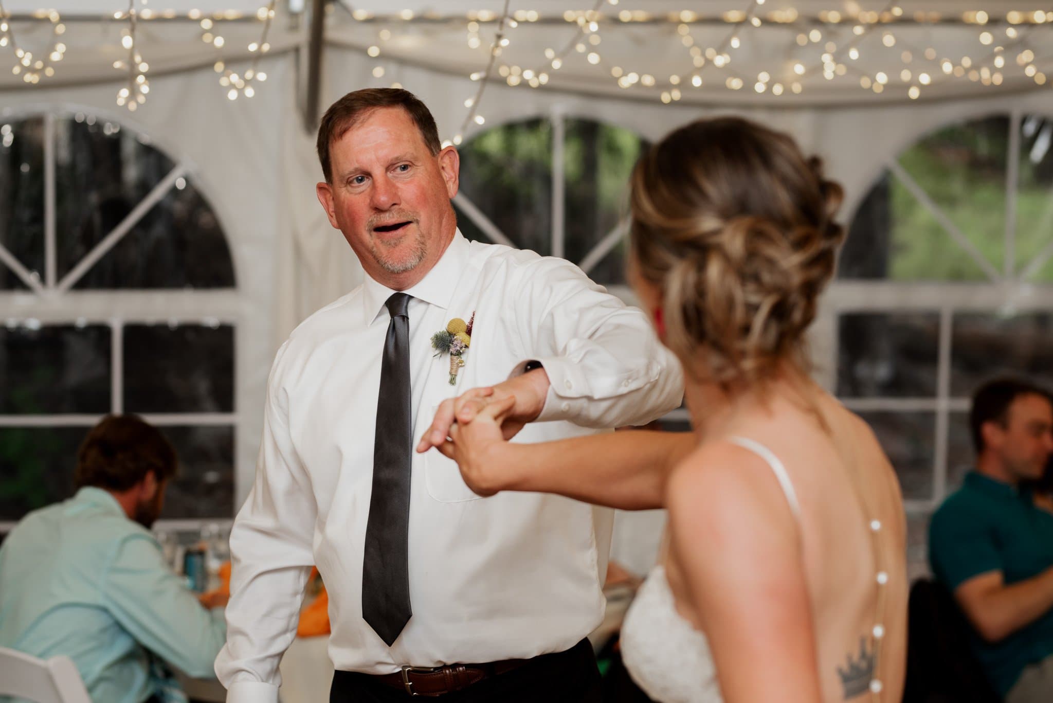A man in a white shirt and black tie shares a joyous dance with a woman in a white dress at what appears to be the father of the bride dance. The venue, Mt Hood Skibowl, is elegantly lit with string lights, and large windows frame the view behind seated guests.
