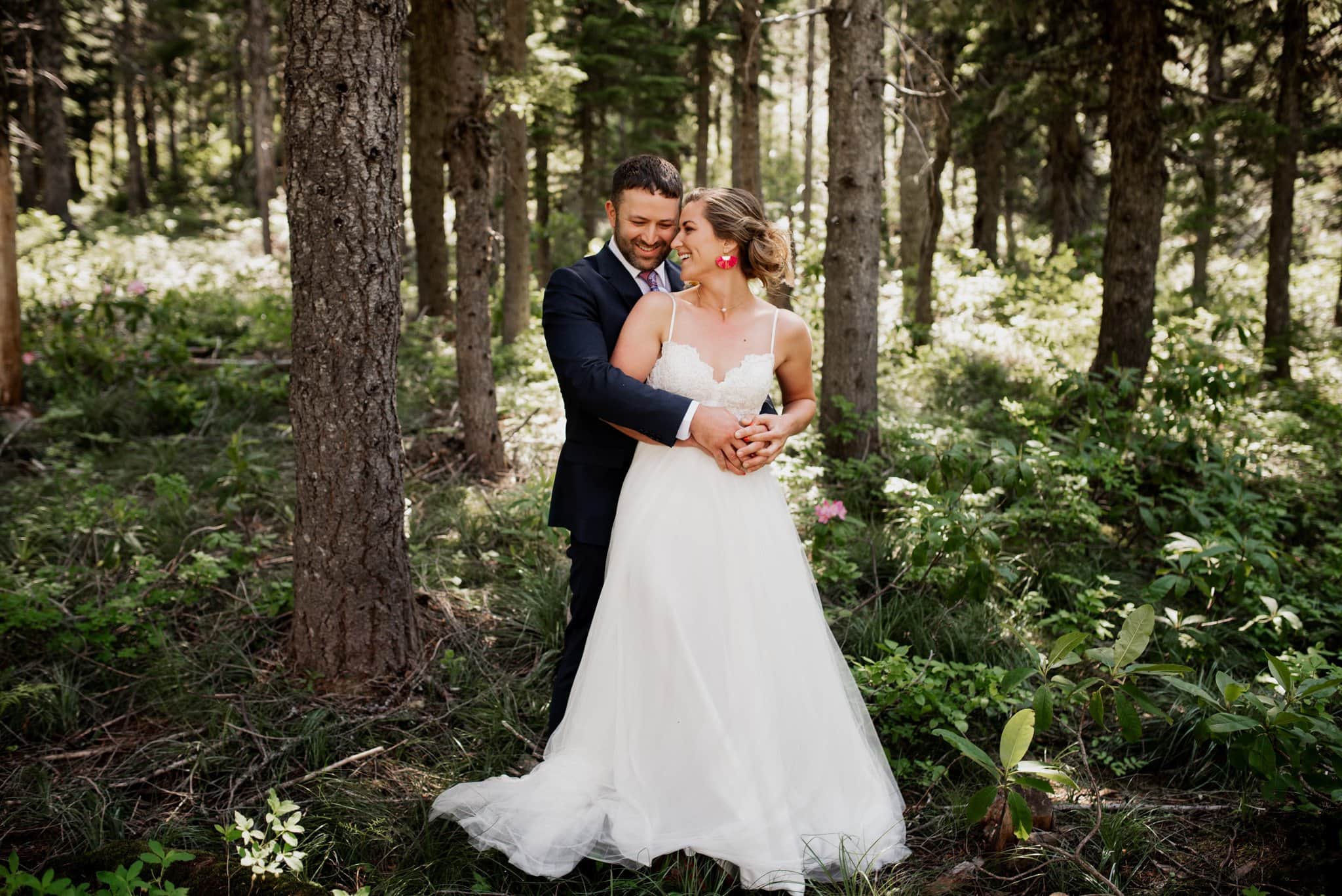 A bride in a white dress and a groom in a dark suit embrace lovingly in a sun-dappled forest at Mt Hood Ski Bowl. The ground is lush with green foliage, creating a serene and romantic atmosphere.