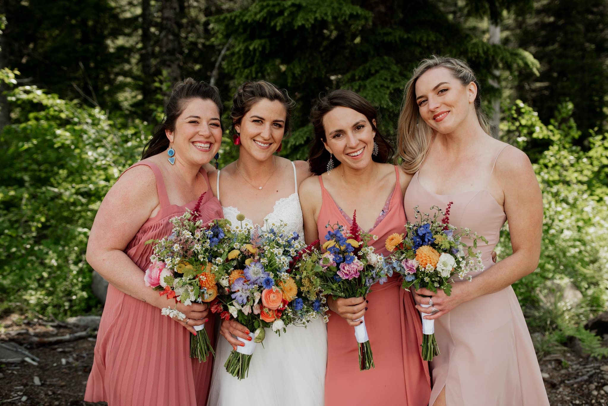 Four women stand together outdoors, each holding colorful bouquets, celebrating at a Mt Hood Ski Bowl wedding. They are dressed in light, summery dresses in shades of pink and white. The background is lush with green trees and foliage. They are smiling and appear joyful.