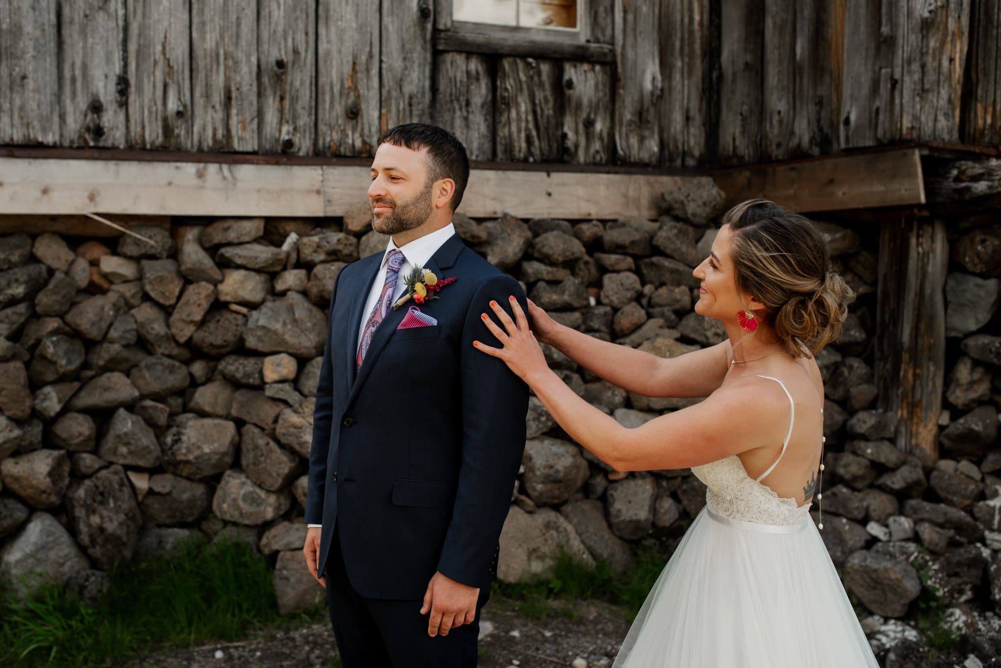In front of a rustic stone and wooden warming hut, a bride in a white dress and red earrings playfully covers the eyes of her groom, echoing their shared joy from the day they met at Mt Hood Ski Bowl. Smiles light up their faces, as vibrant as their cherished memories.