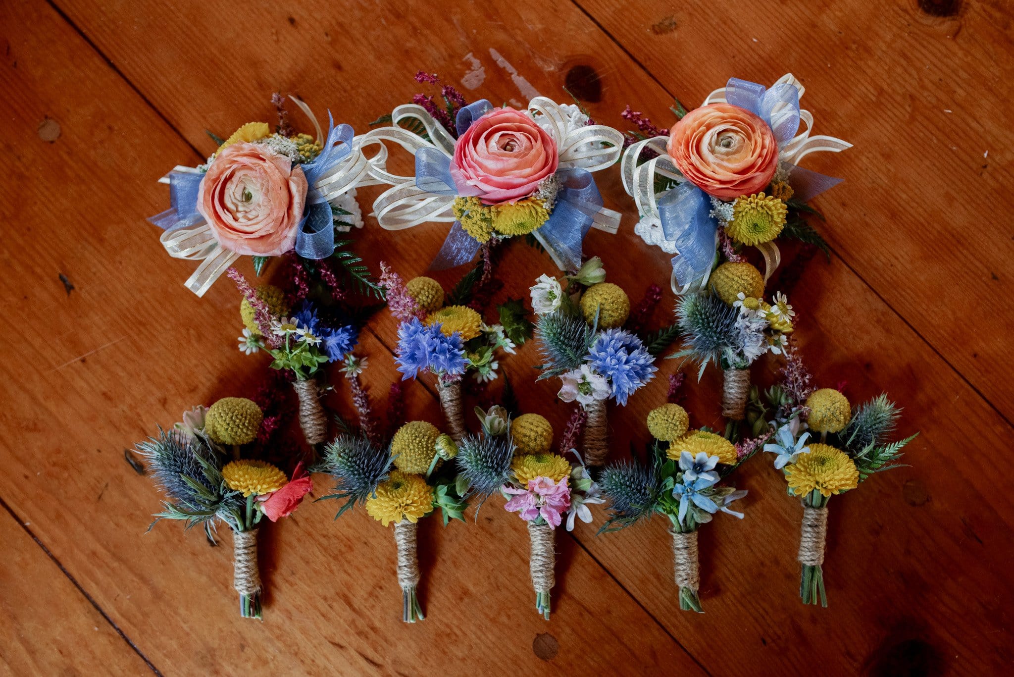 A group of colorful boutonnieres and corsages, perfect for a Mt Hood Ski Bowl wedding, arranged neatly on a wooden surface. Each piece features pink roses, blue thistles, yellow billy balls, and greenery, tied with twine and some adorned with ribbon bows.