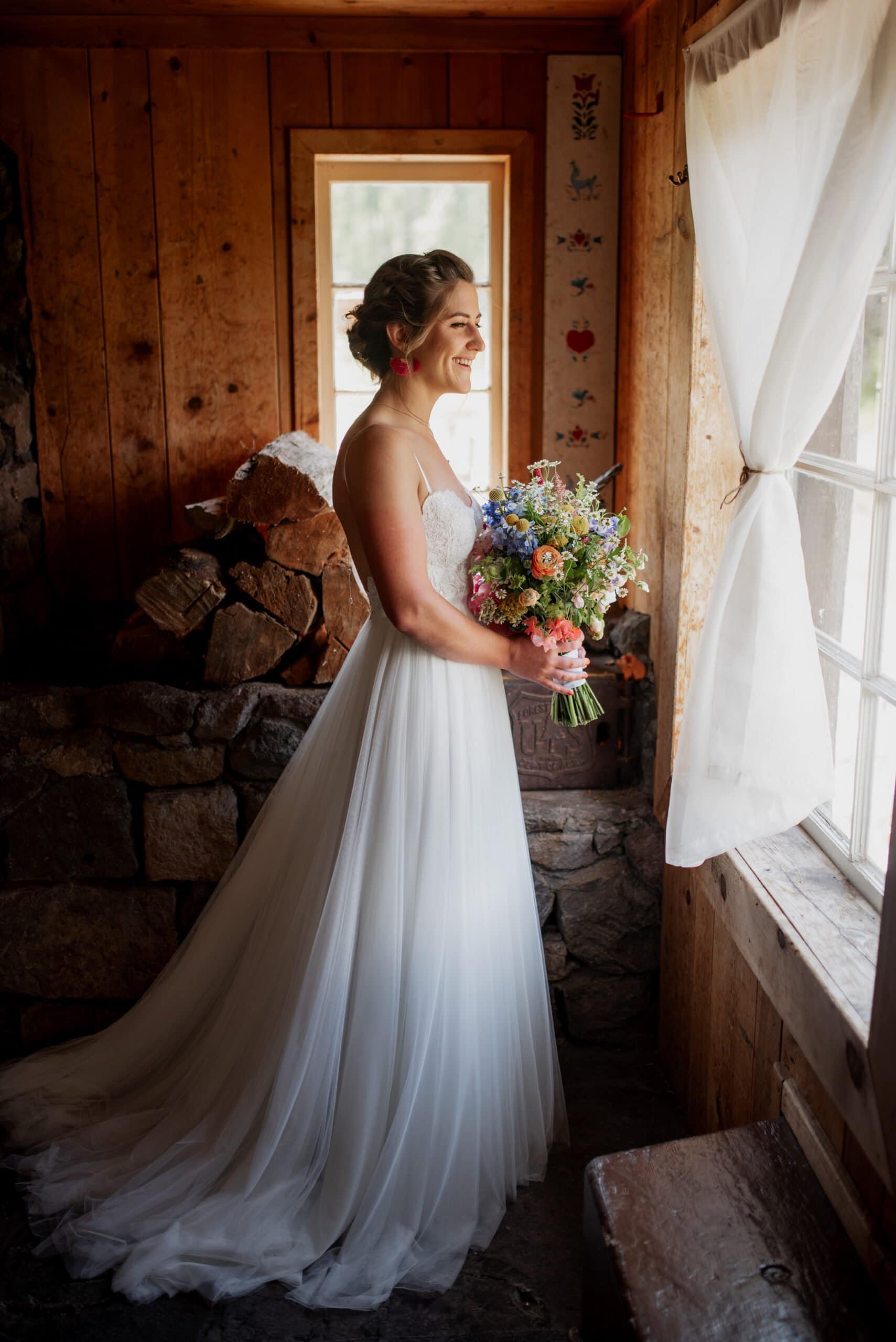 A bride in a flowing white gown, holding a colorful bouquet, gazes out a sunlit window in the Mt Hood Skibowl warming hut. She stands in a rustic wooden warming hut with stone details, her hair styled elegantly. Bright light filters through the window, creating a warm, serene atmosphere.