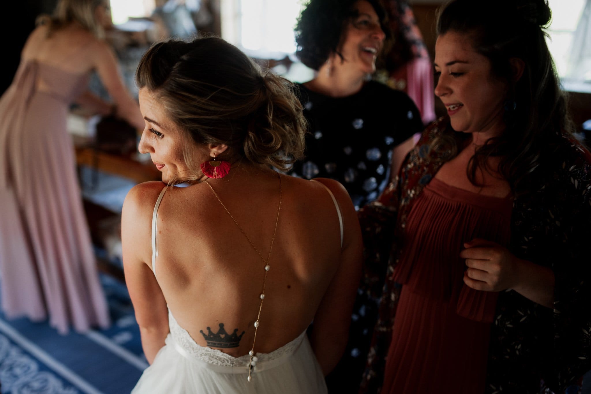 A bride with a crown tattoo on her back smiles at two women. One wears a black dress with white dots, the other a red dress. A woman in a pink dress is in the background. The joyful atmosphere of the Mt Hood Ski Bowl wedding is accentuated by the dim lighting.