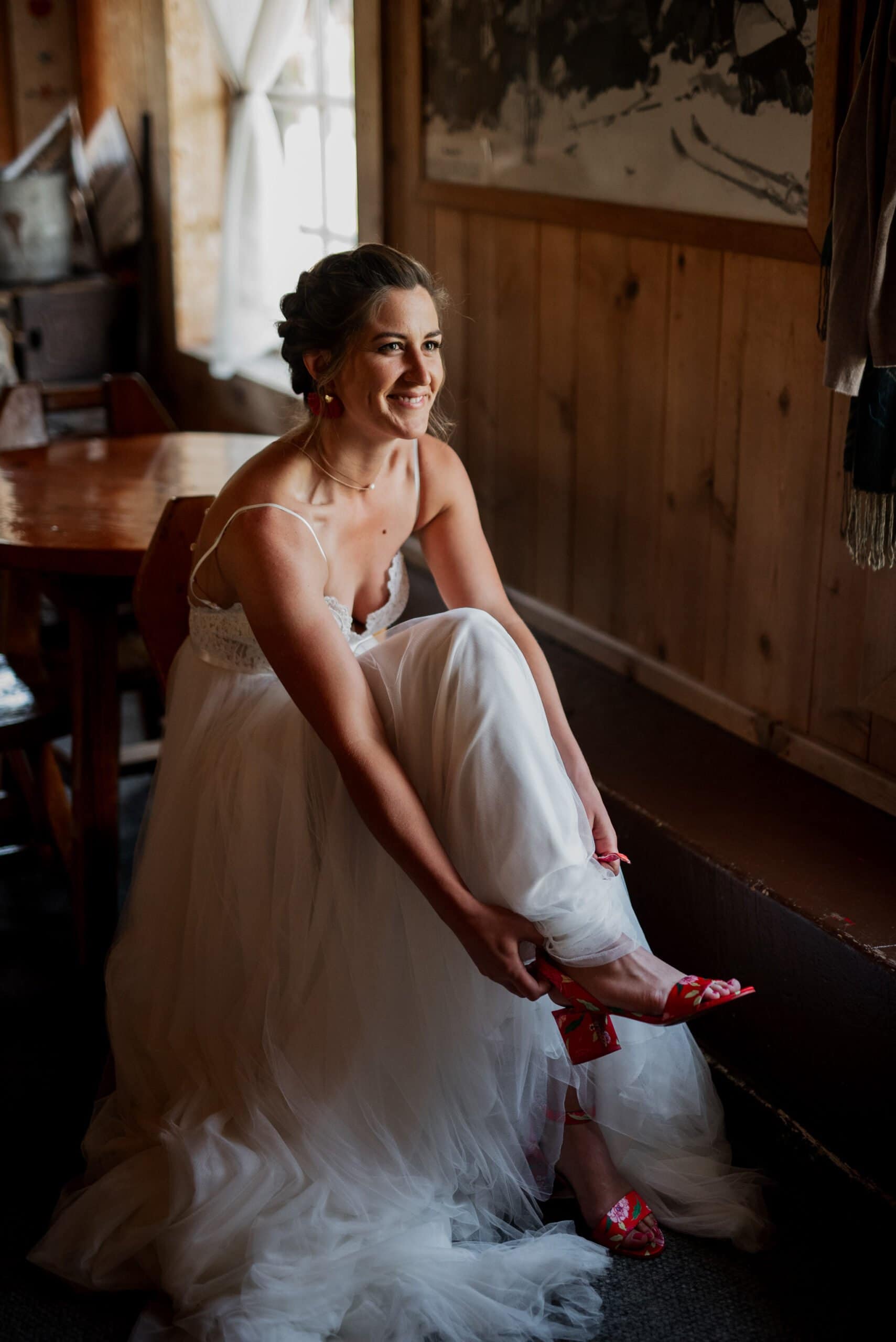 A bride in a white wedding gown sits on a wooden bench, smiling and adjusting her red shoes with floral patterns. The cozy room, resembling a Mt Hood Ski Bowl lodge, features wooden walls and furniture as soft light filters through the window.