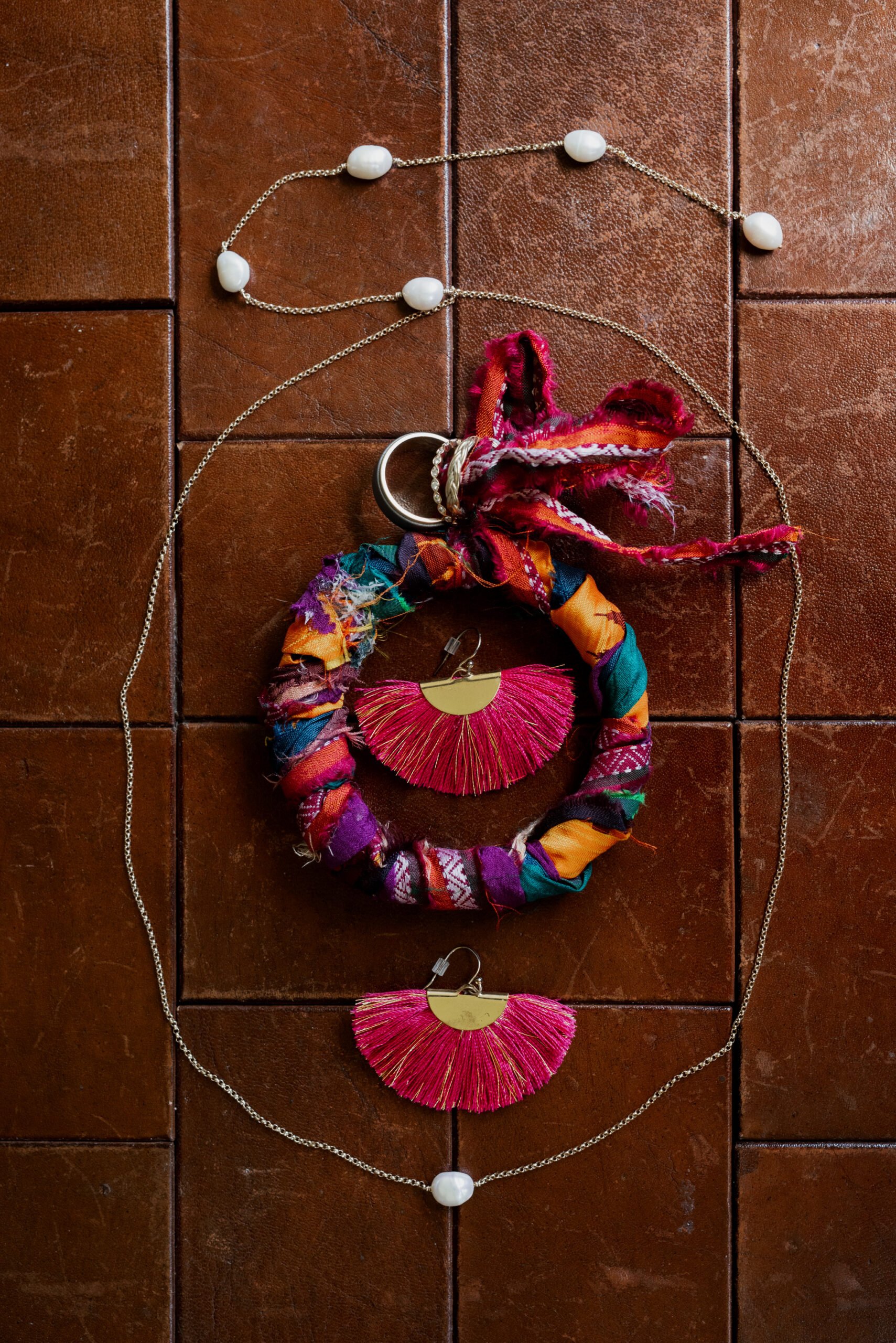 A colorful textile bracelet with a silver ring and red tassels joins red tassel earrings and a gold chain necklace with white beads, elegantly arranged on a brown textured surface—perfect accessories for a Mt. Hood Ski Bowl wedding.