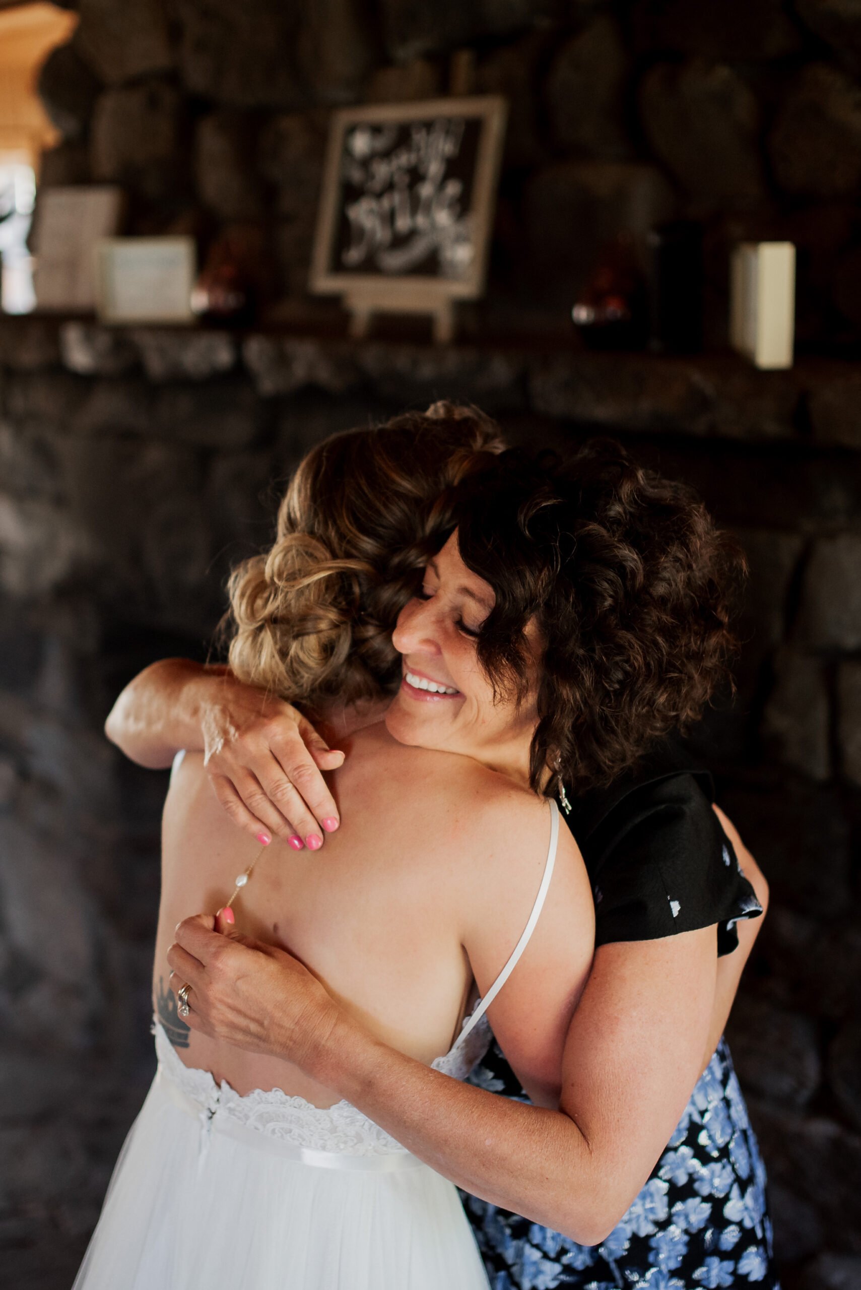 Two women share a warm embrace in front of a stone wall at a Mt. Hood Ski Bowl wedding. One is in a white dress, the other in a floral-patterned outfit, both radiating happiness. The woman in the floral dress smiles widely as a framed picture graces the mantle behind them.