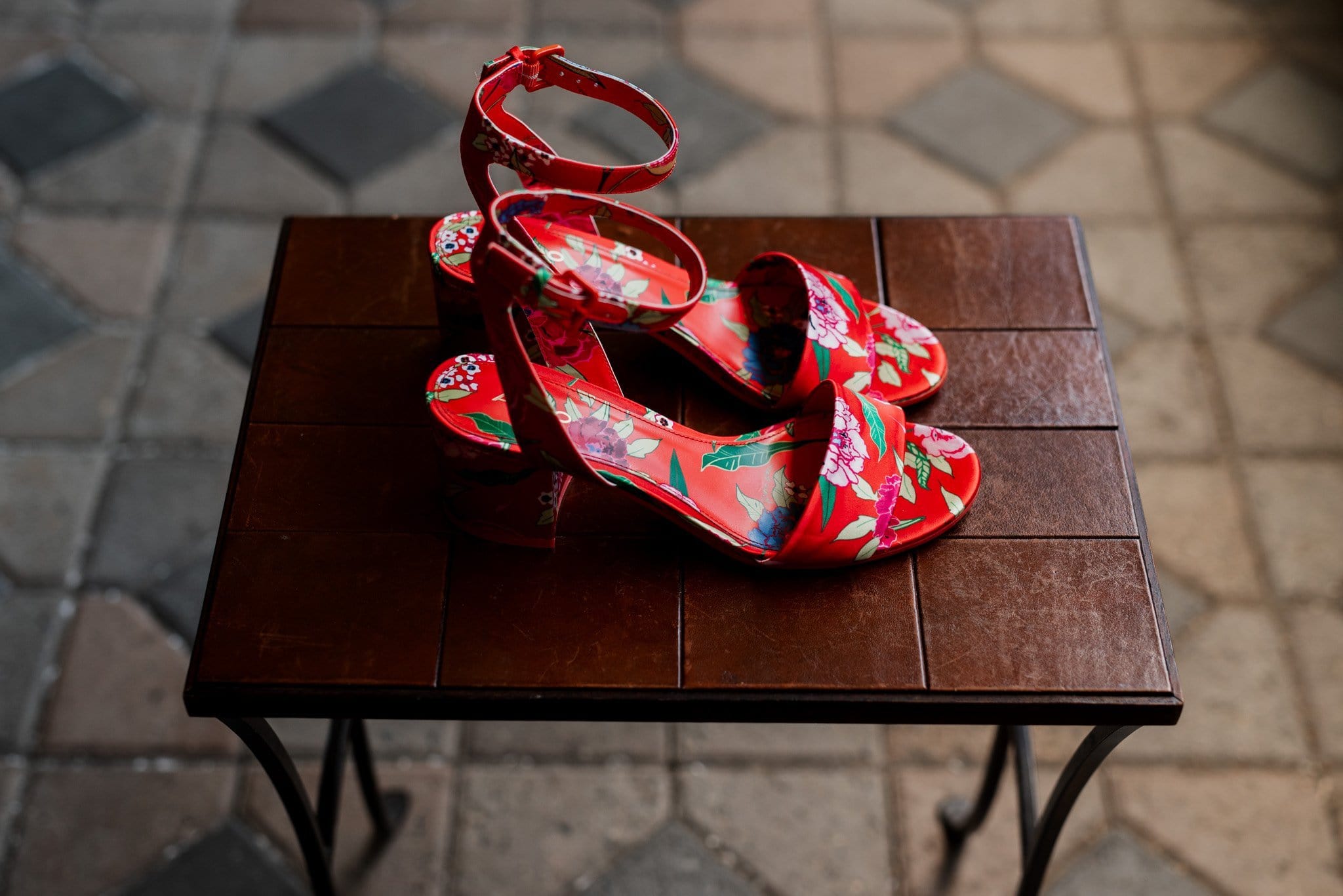A pair of vibrant red floral high-heeled sandals with ankle straps rest gracefully on a small wooden table, set against a patterned tiled floor. Perfect for a Mt Hood Ski Bowl wedding, the sandals boast a summery, elegant design that captures attention.