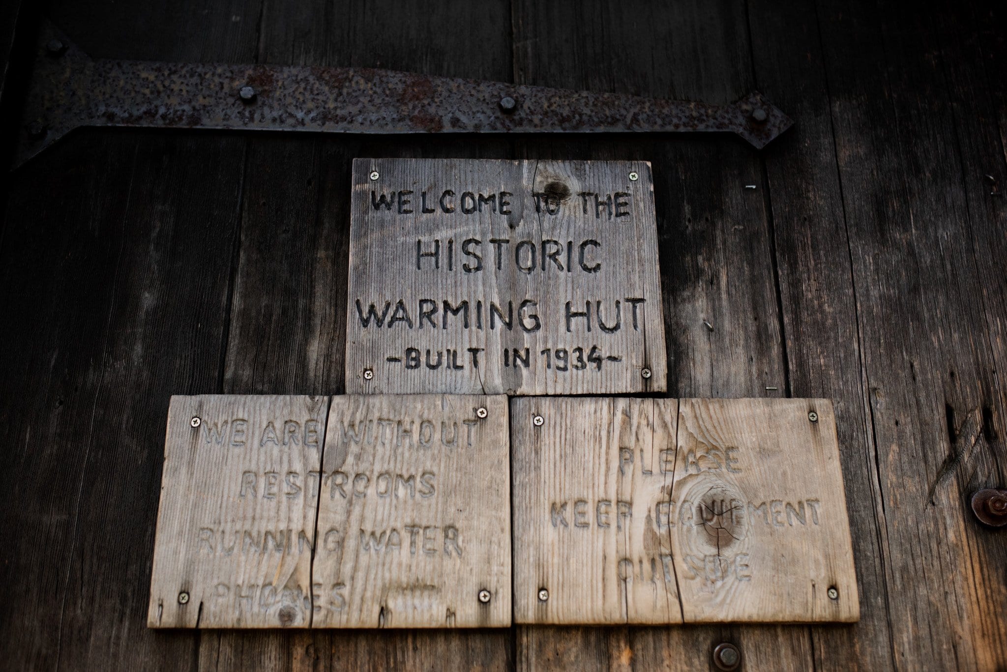 Weathered wooden signs on a wall read: Welcome to the Historic Warming Hut - Built in 1934, We are without restrooms running water phones, and Please keep equipment outside. Ideal for an intimate Mt Hood Ski Bowl wedding backdrop.