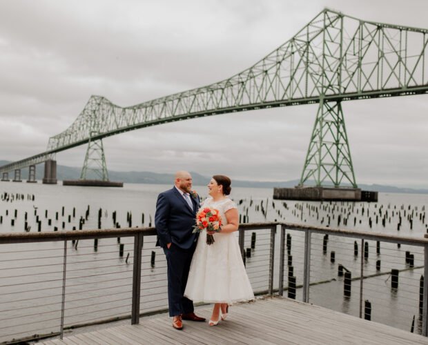 A couple in wedding attire poses on a pier with a large bridge in the background under a cloudy sky, capturing memorable photos on their special day at The Loft at the Red Building.
