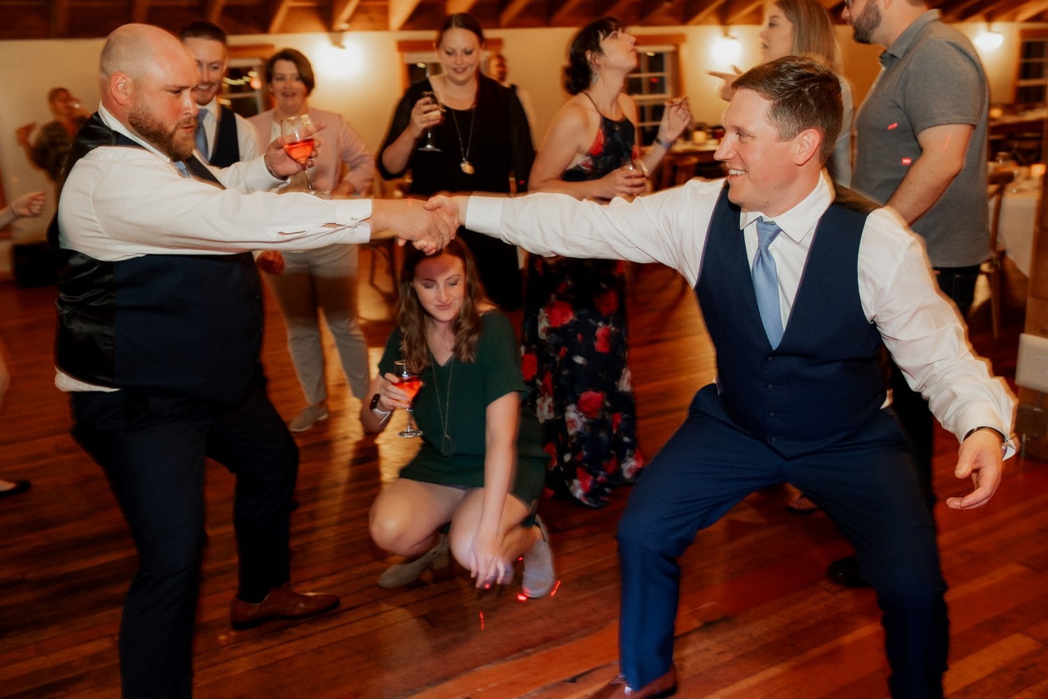 A lively party scene with guests dancing energetically. One man in a blue suit is playfully clapping or interacting with another guest. This moment is captured during a wedding celebration at The Loft at the Red Building.