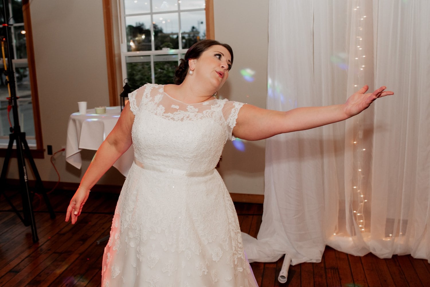 The bride, wearing a beautiful white gown, joyfully dances with one arm extended amidst the festive decor at her wedding at The Loft at the Red Building.