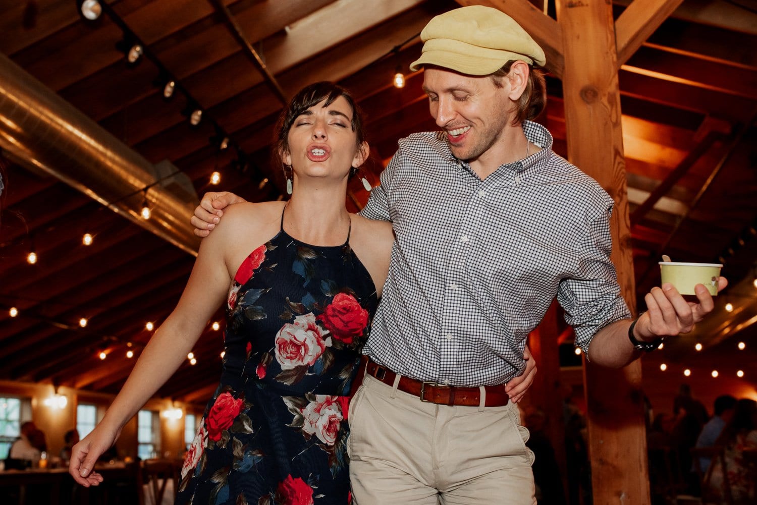 A woman and man casually enjoy a moment together in a warmly lit rustic venue, with brick walls and wooden accents visible in the background. The warm lighting creates a cozy atmosphere as the couple stands close to each other, smiling and appearing relaxed.